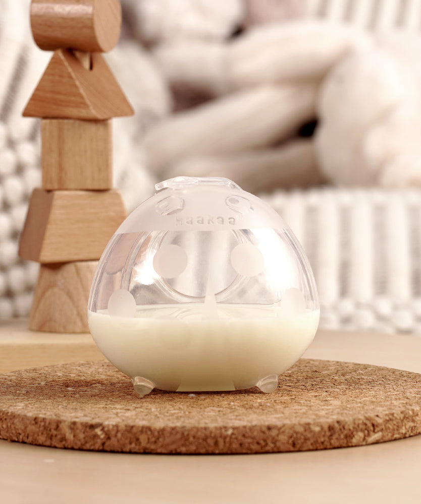 The Haakaa Ladybug Silicone Breast Milk Collector, filled with milk is stood on a light brown table and is stable due to the little silicone feet on the bottom of the milk collector