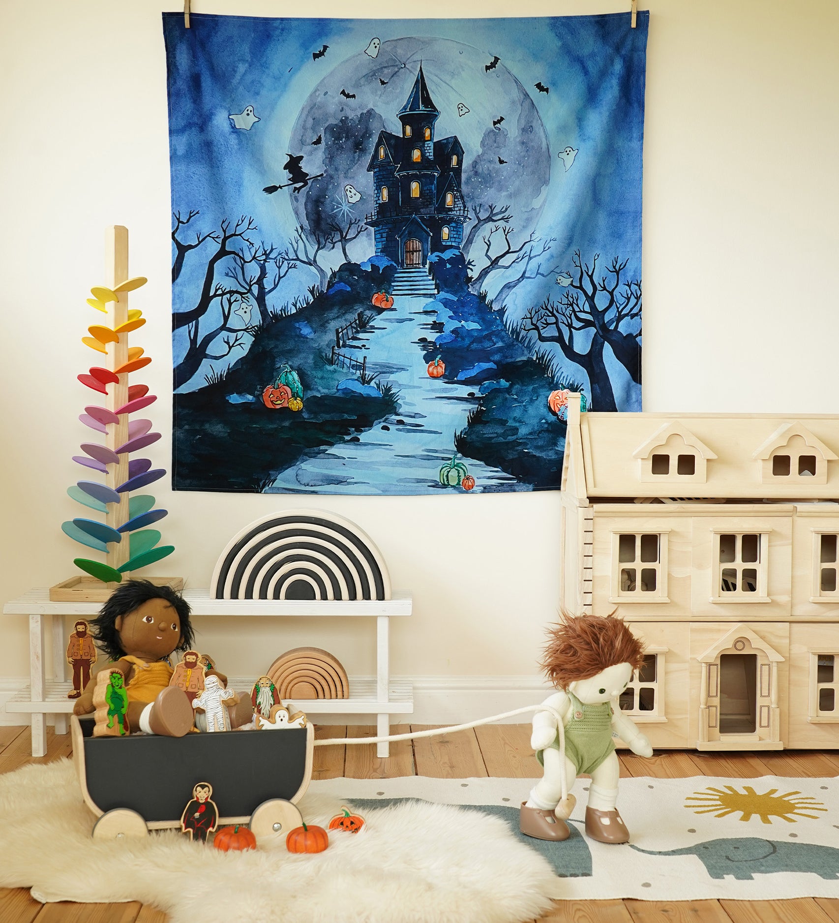 The Wonderie Play Cloth - Spooky Manor hanging up on a wall in a play room. The image shows Halloween toys, a dolls house, a Grimm's rainbow and a marble tree 