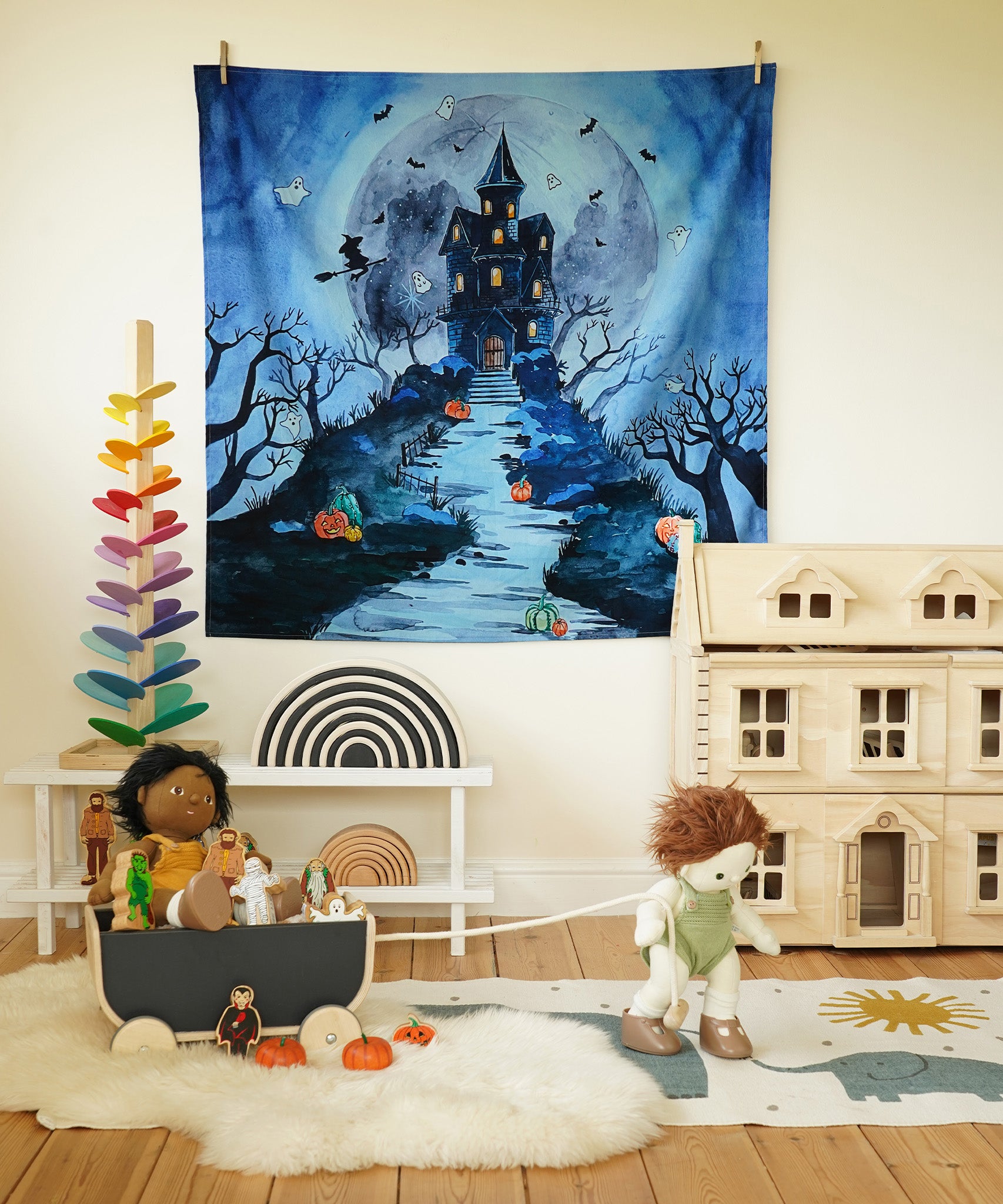 The Wonderie Play Cloth - Spooky Manor hanging up on a wall in a play room. The image shows Halloween toys, a dolls house, a Grimm's rainbow and a marble tree 