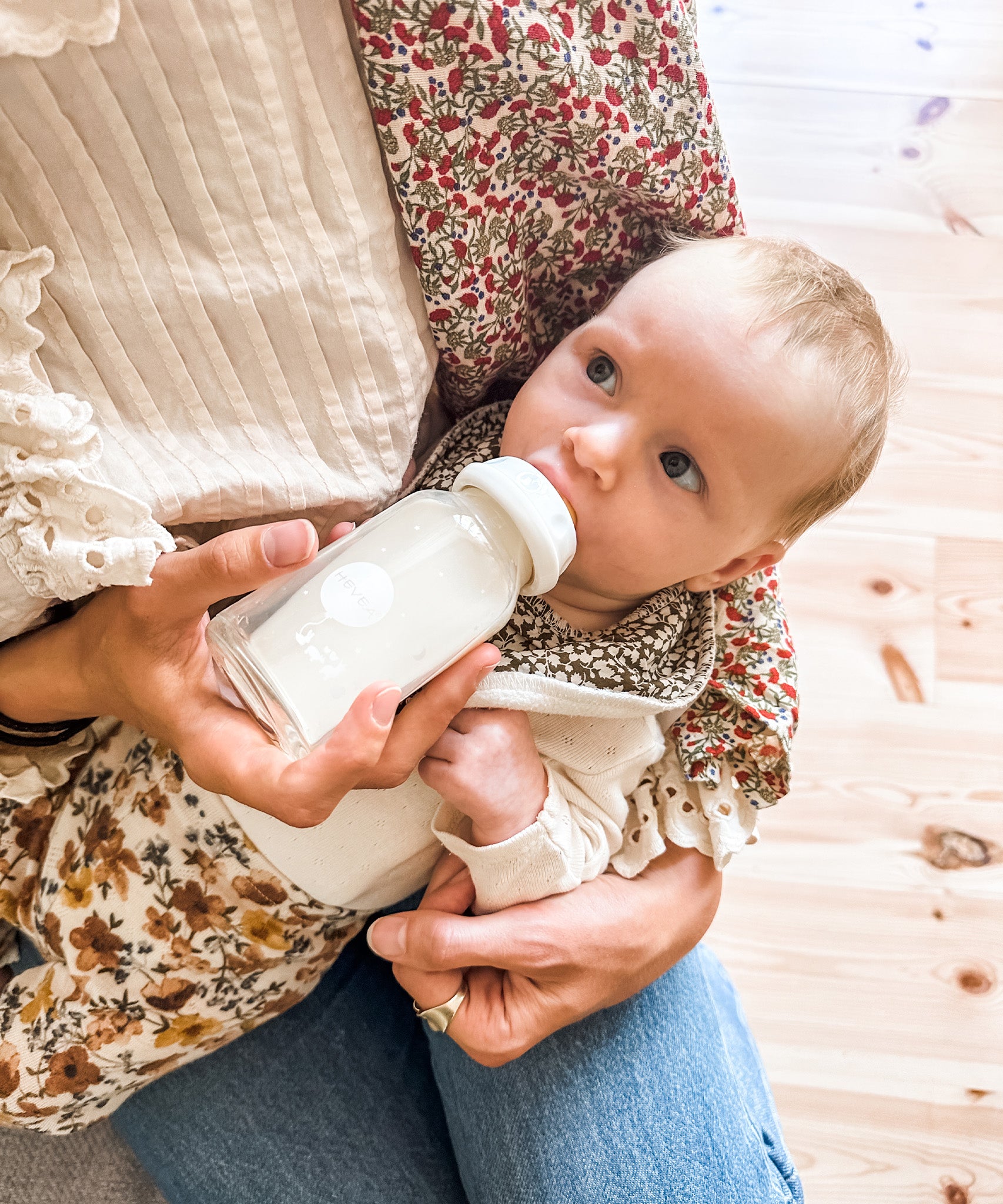 Baby feeding from a Hevea glass baby bottle on parents lap.