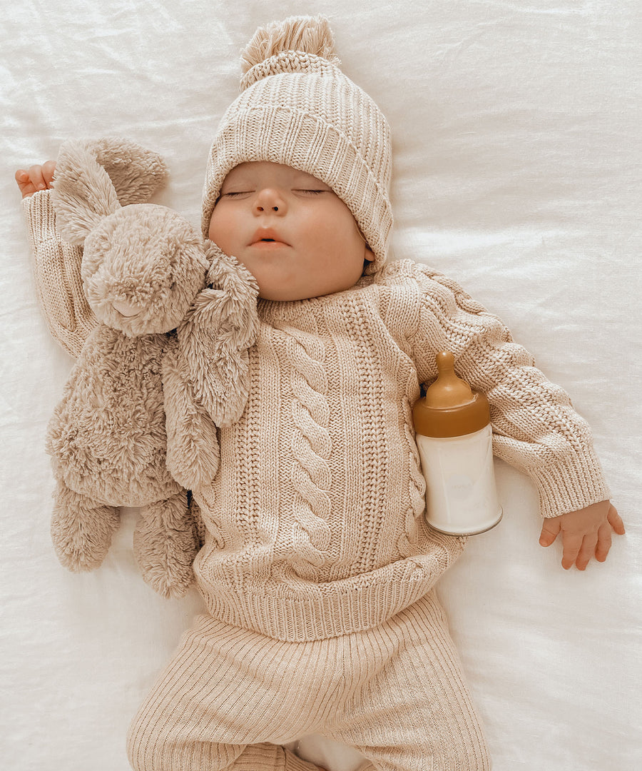 Baby wearing a cream knitted hat and jumper asleep with a Hevea Glass Baby Bottle and toy rabbit.