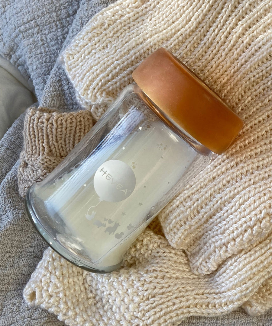 Hevea Wide Neck Glass Baby Bottle with Natural Rubber Cap on a white knitted blanket. 