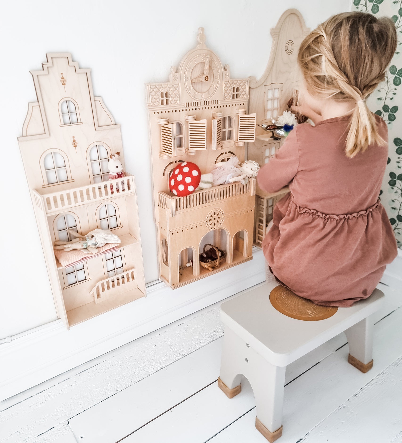 Child in a pink dress sat on a Hevea Rubberwood Step Stool, playing with a dolls house.