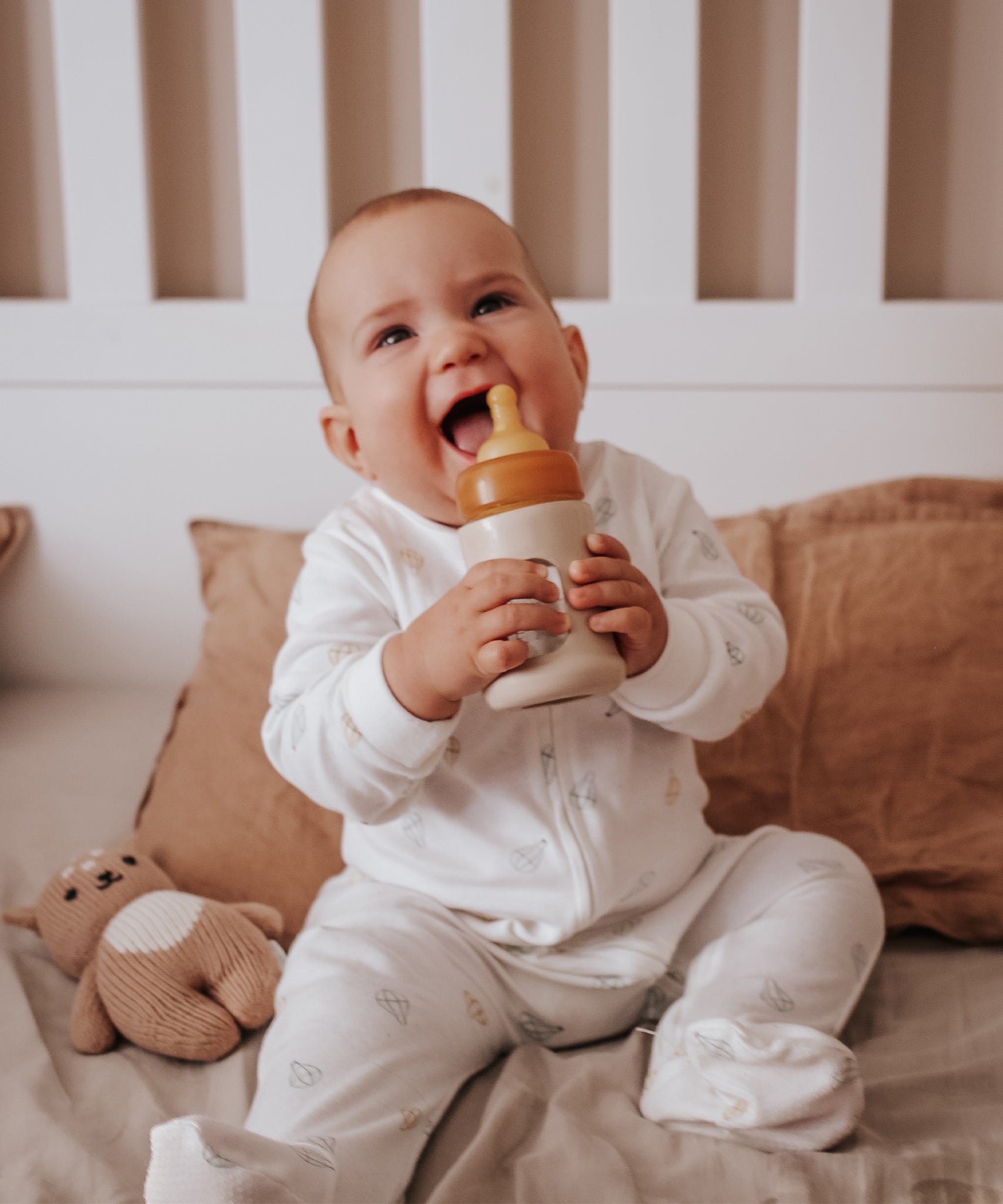 Baby sat in a cot smiling with the Hevea Glass Baby Bottle Wide Neck With Rubber Sleeve - 150ml in Sand.