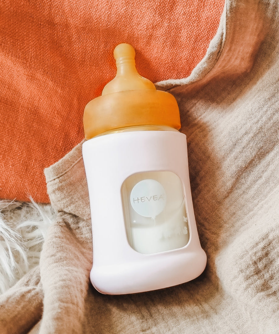 Hevea Glass Baby Bottle Wide Neck With Rubber Sleeve - 150ml in Sand on cream and terracotta muslins.