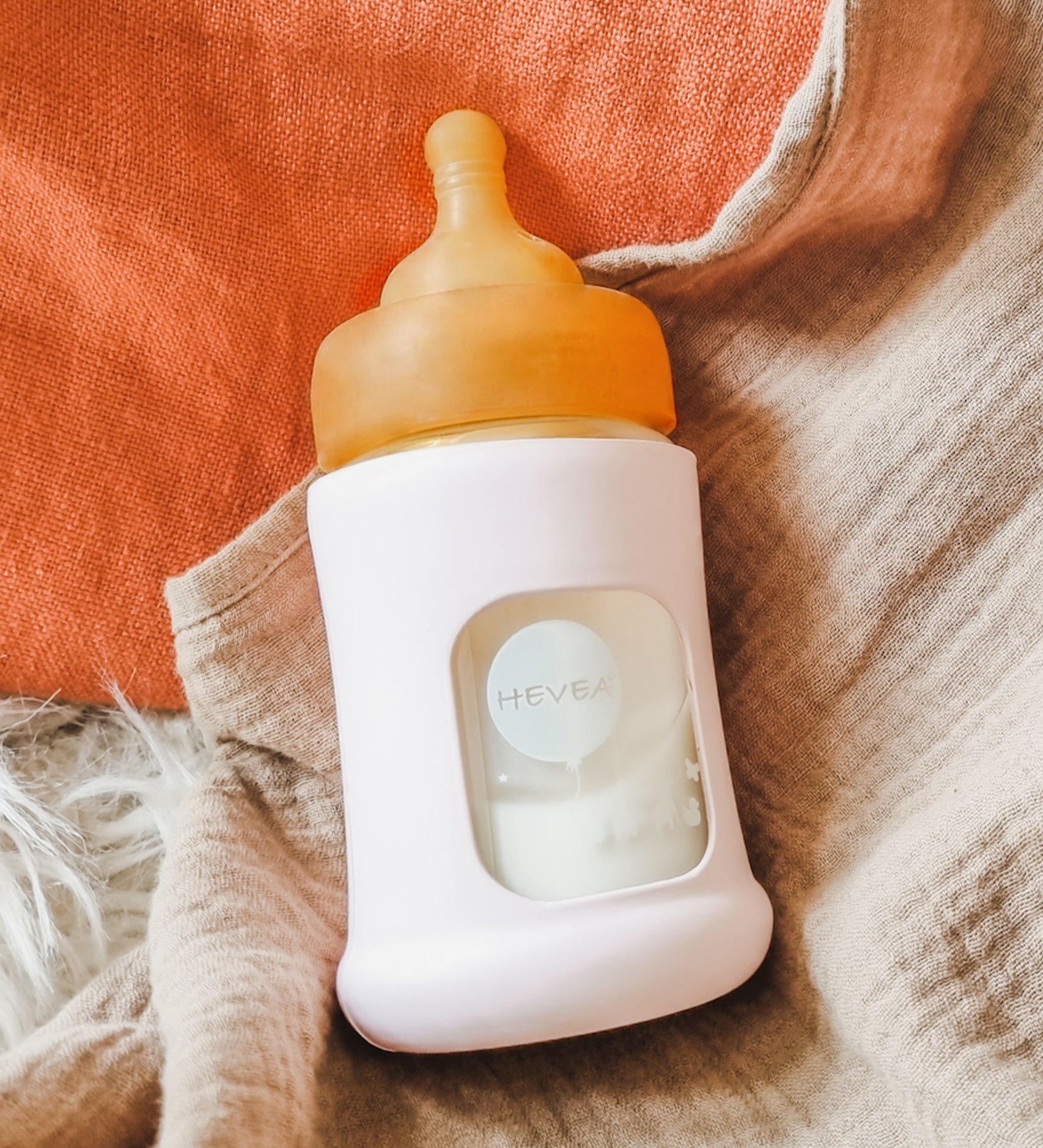 Hevea Glass Baby Bottle Wide Neck With Rubber Sleeve - 150ml in Sand on cream and terracotta muslins.