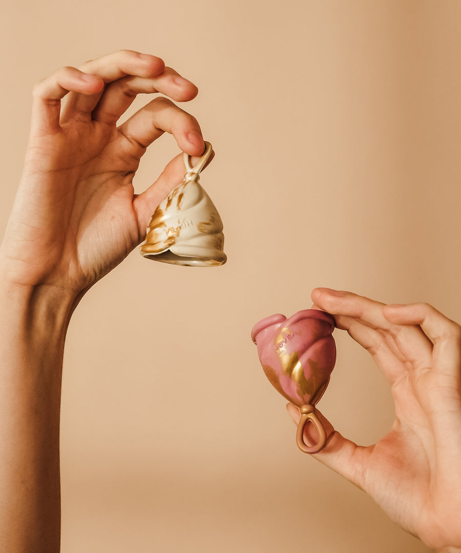 Person holding up two menstrual cups, one cream and gold, the other pink and gold.