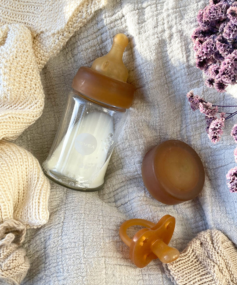 A Hevea Glass Baby Bottle half filled with milk, a natural rubber cap and dummy on a white muslin.