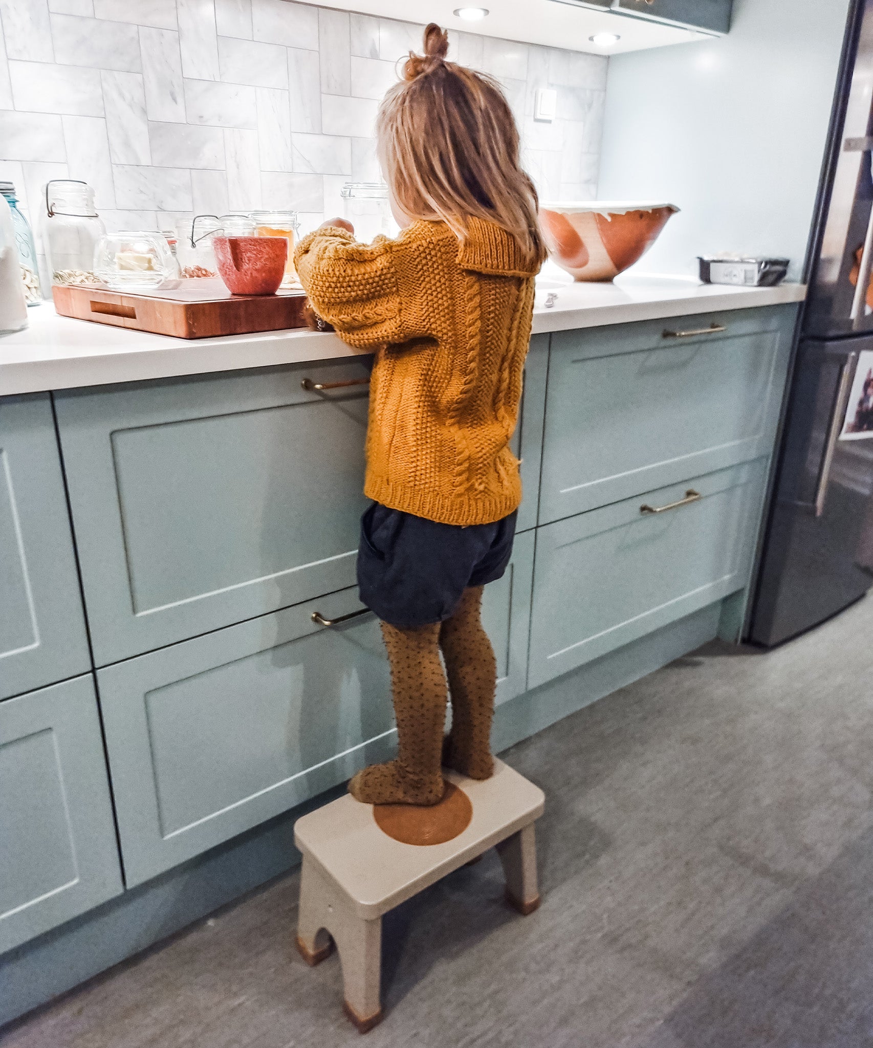 Child in yellow tights preparing food stood on a Hevea Rubberwood Step Stool at a kitchen counter.