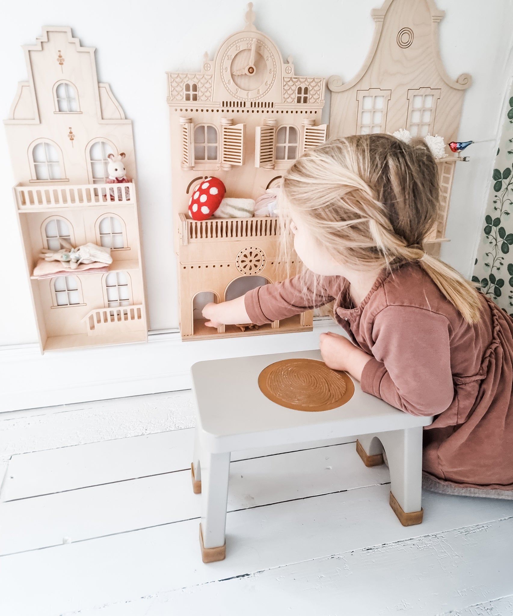 Child in a pink dress sat on a Hevea Rubberwood Step Stool, playing with a dolls house.