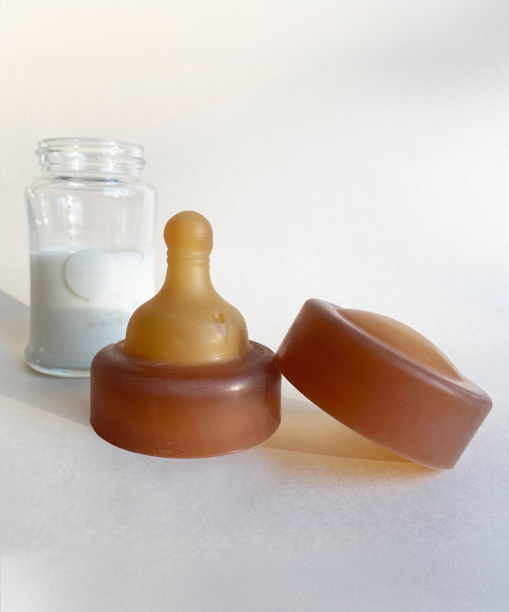 A Hevea Glass Baby Bottle with milk in the background with the natural rubber teat and cap in the foreground. 