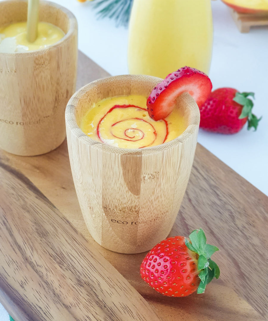 A yellow mango smoothie with red strawberry puree swirl in a bamboo baby cup, decorated with strawberry slices..