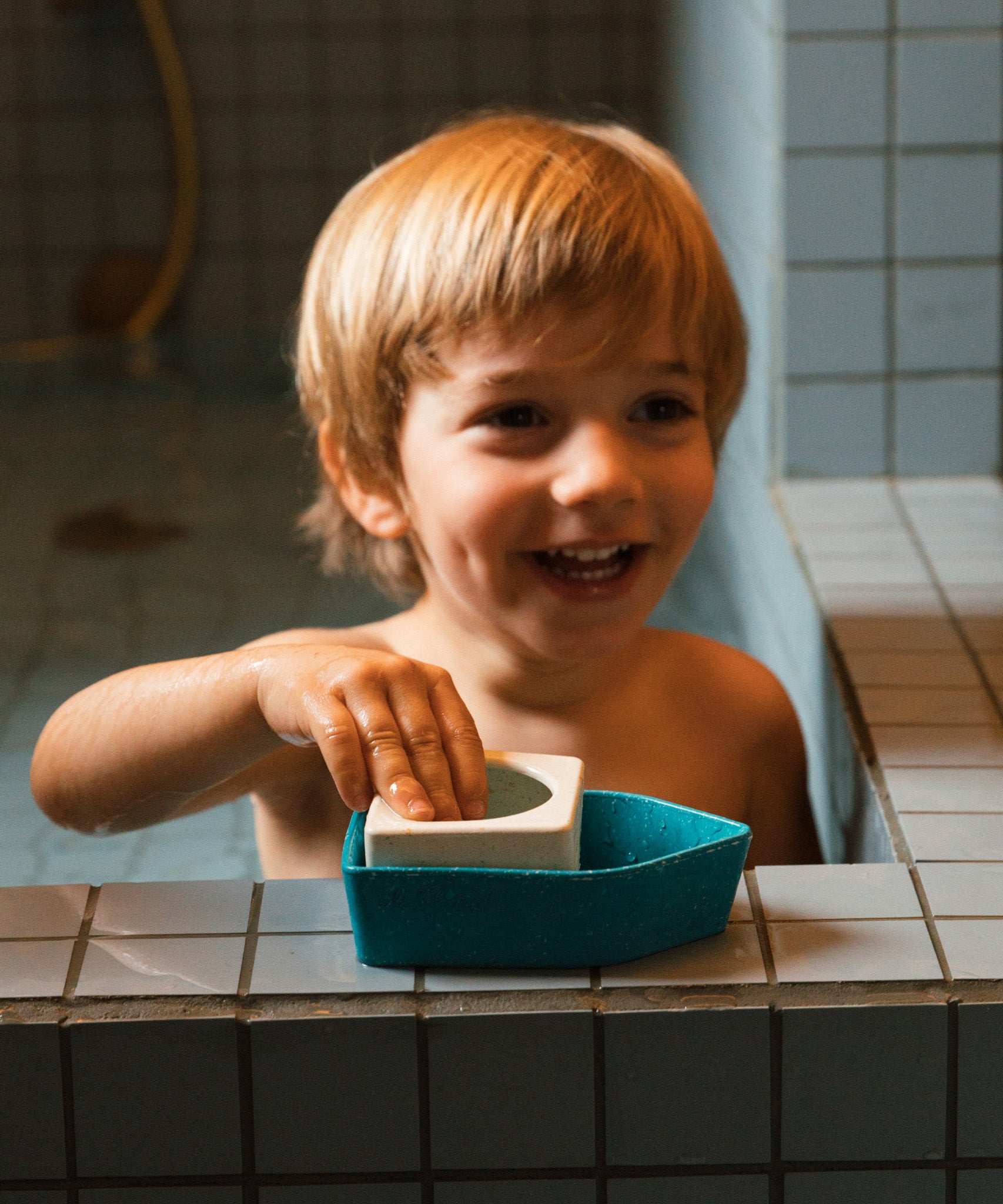 A child playing with the Oli & Carol Boat Bath Toy - Yellow, on the side of a blue tiled bath