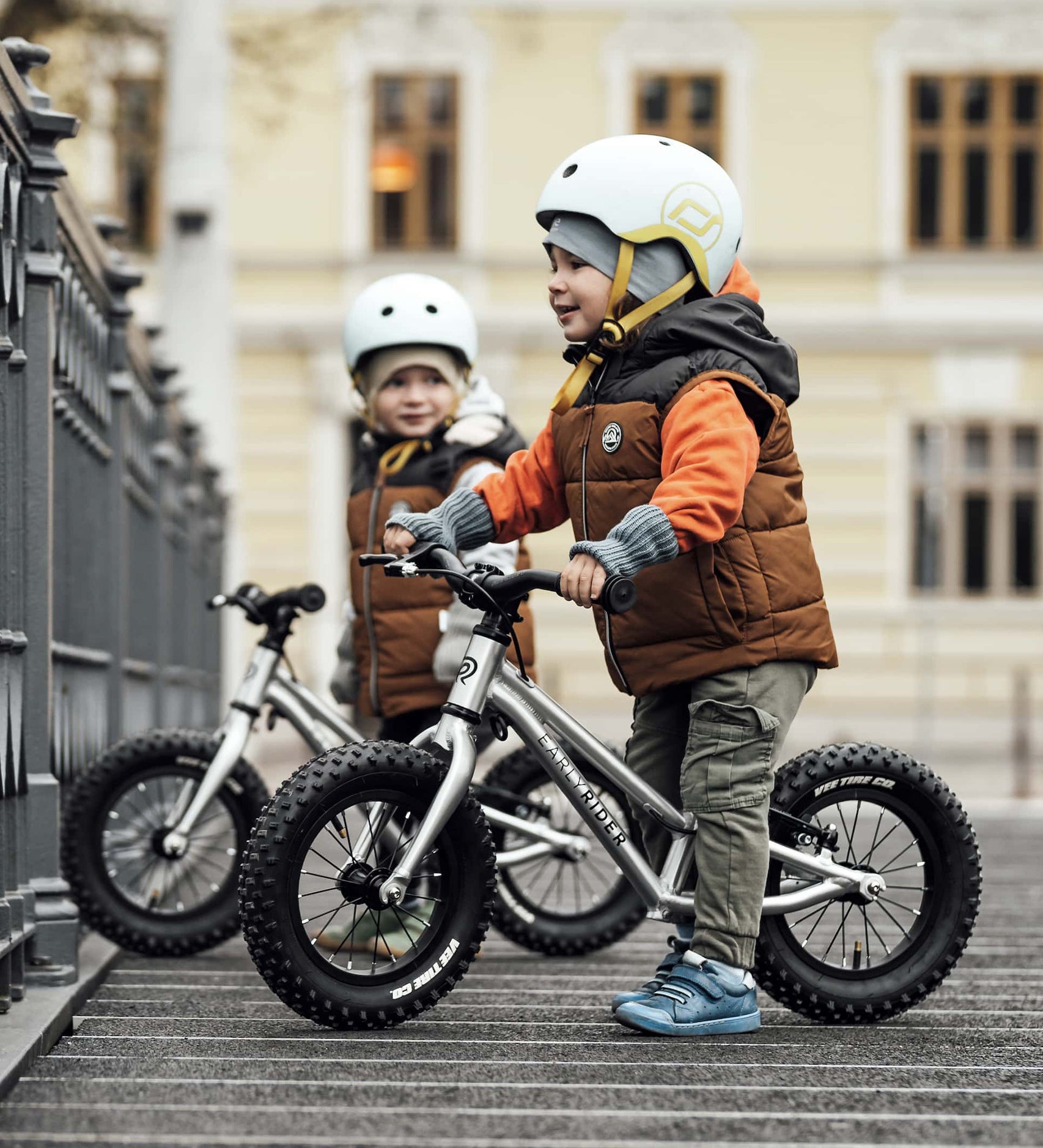 Two children happily stood with their Early Rider Big Foot Balance Bikes next to a railing, with a large light yellow building in the background