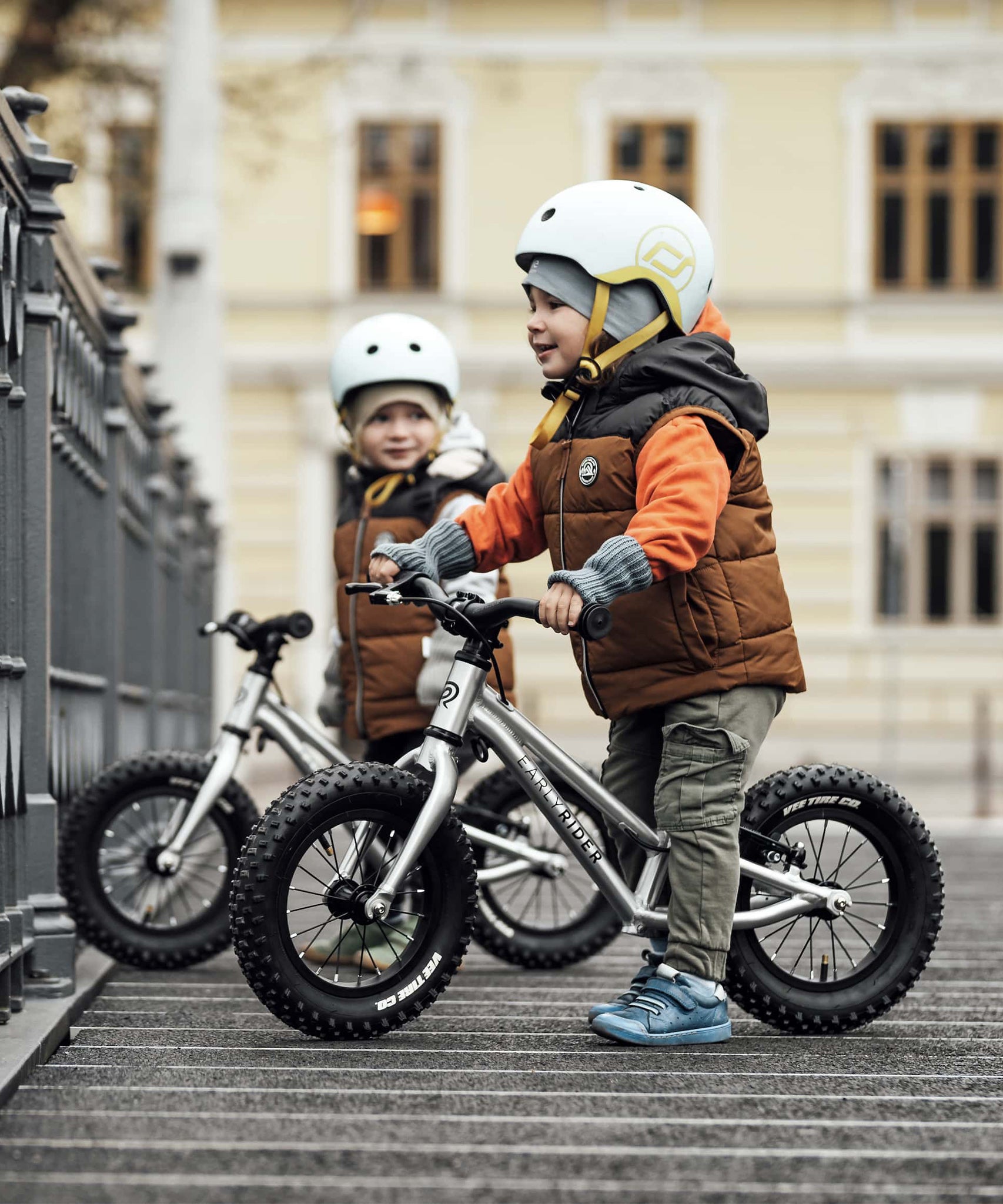 Two children happily stood with their Early Rider Big Foot Balance Bikes next to a railing, with a large light yellow building in the background