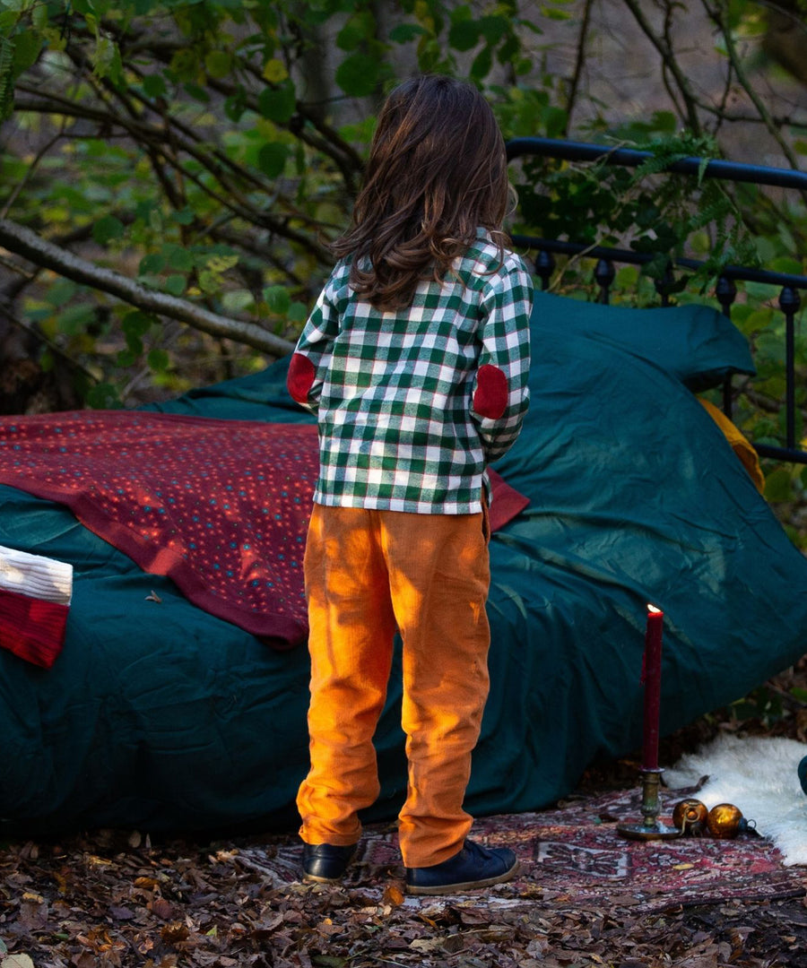 A child stood next to a bed with green bedding, outside in a forest. The child is wearing the Little Green Radicals Check Long Sleeve Shirt - Fern Green, and showing the red elbow patches. The child is also wearing yellow trousers