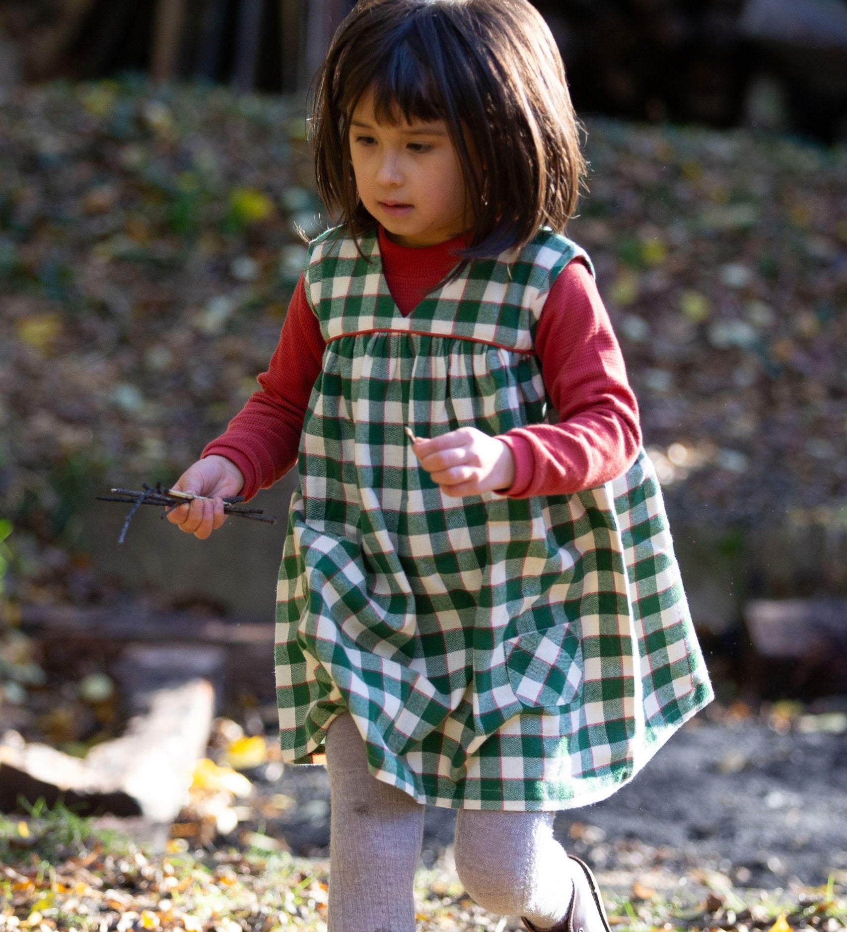 A child outside, playing in the sun and collecting sticks, wearing the Little Green Radicals Long Sleeve Waffle Top - Hazelnut underneath a green check pinafore dress