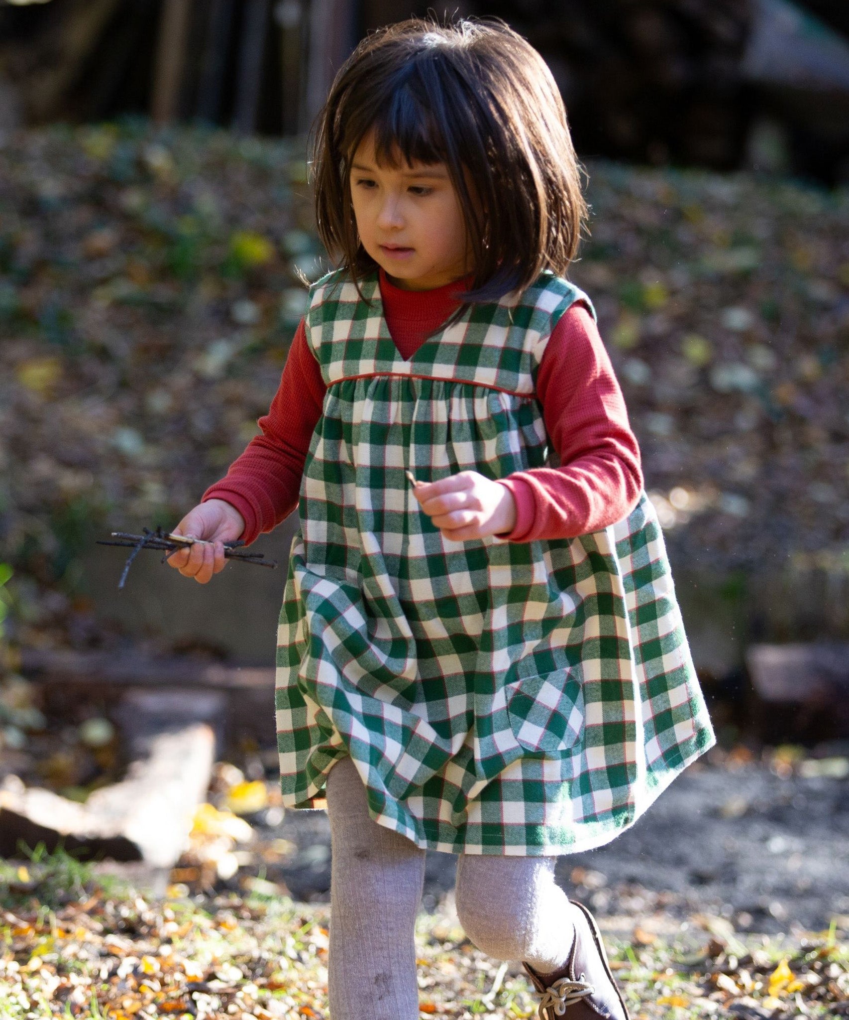 A child outside, playing in the sun and collecting sticks, wearing the Little Green Radicals Long Sleeve Waffle Top - Hazelnut underneath a green check pinafore dress