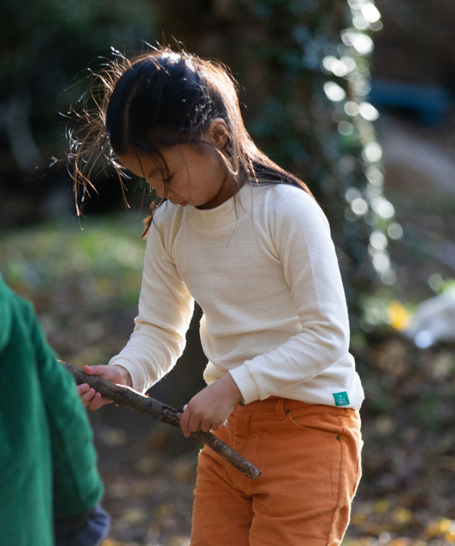 A child is looking curious about the stick they are holding in their hand. They are wearing the  Little Green Radicals Long Sleeve Waffle Top - Cream, with yellow trousers