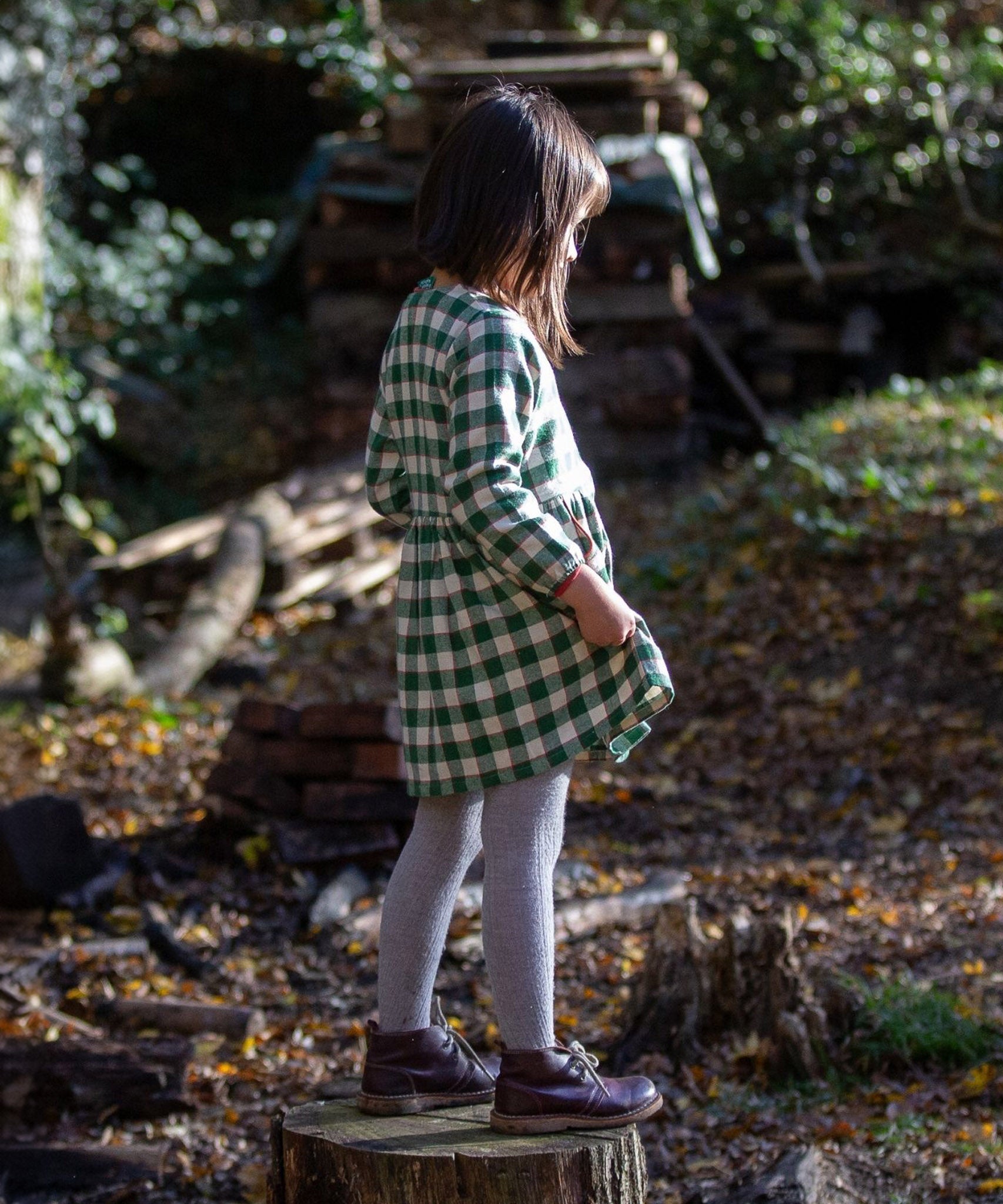 A child is stood on a wooden tree stump, wearing the Little Green Radicals Check Pocket Dress - Fern Green. The image shows the child from the side