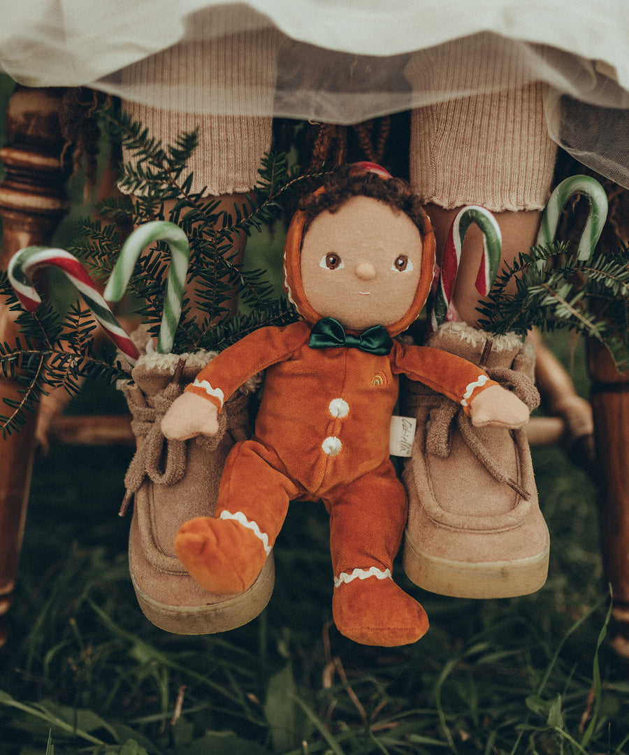 Georgie Gingerbread is sat between a child's feet. The child is wearing tan/pink boots with candy canes and small pine tree branches poking out to give a festive and Christmas feel.