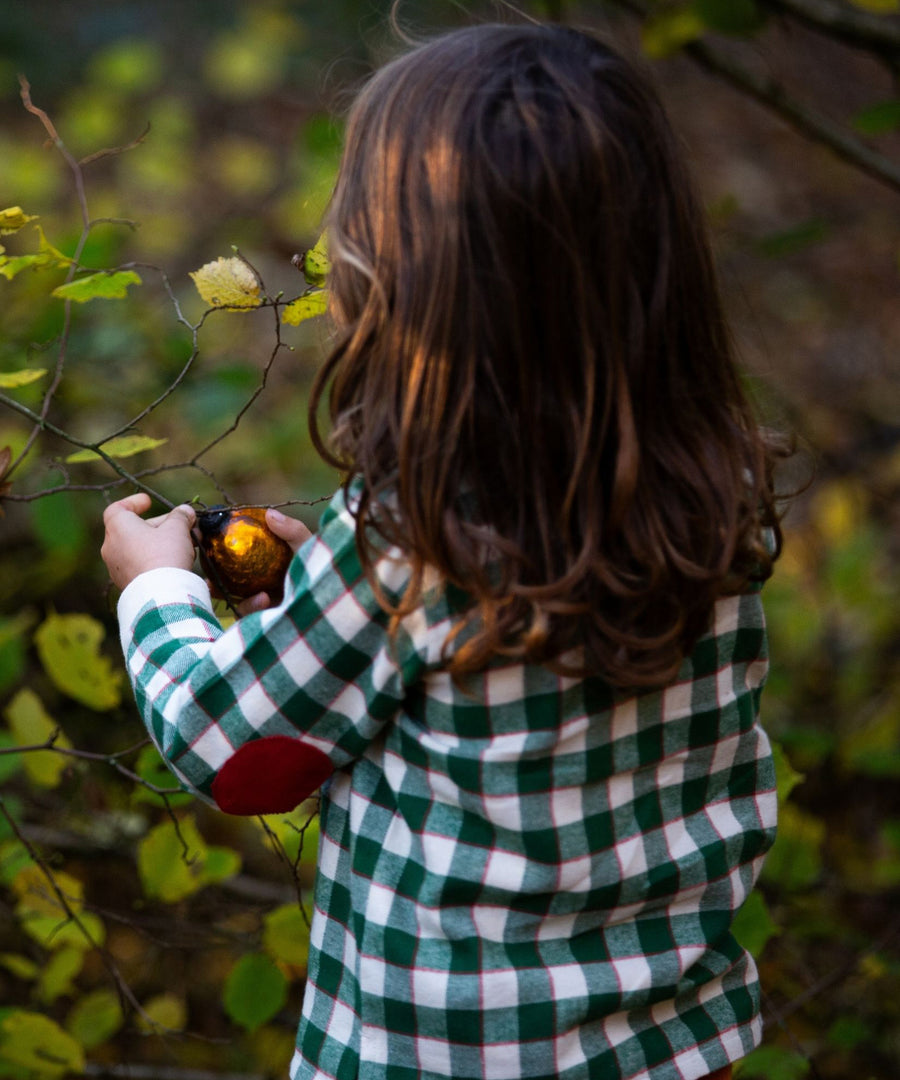 A child hanging decorations onto a tree, wearing the Little Green Radicals Check Long Sleeve Shirt - Fern Green. The image is showing the back of the shirt, and the red elbow patch