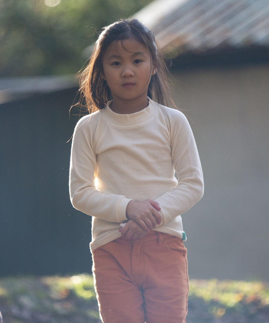 A child facing front, and holding their hands together, wearing the  Little Green Radicals Long Sleeve Waffle Top - Cream