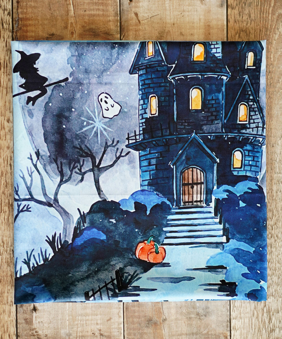 A closer look at the beautiful spooky print on the Wonderie Play Cloth - Spooky Manor. The image is a close up of the manor, showing the brick work and the light on inside. A pumpkin is next to the path, a spooky ghost and witch are flying next to the manor with leafless trees underneath them