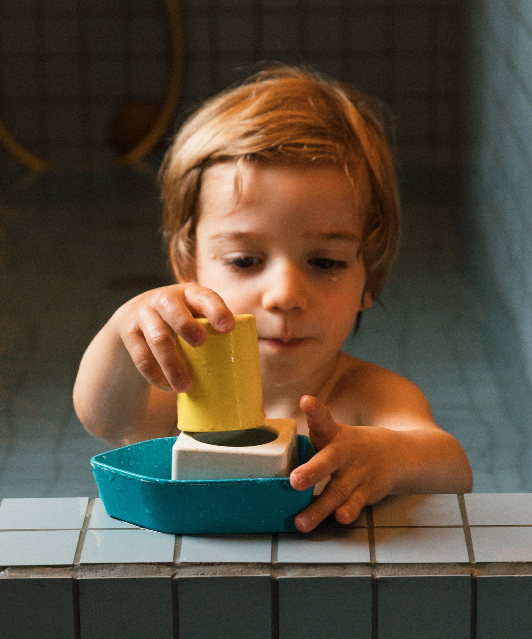 A child playing with the Oli & Carol Boat Bath Toy - Yellow, on the side of a blue tiled bath