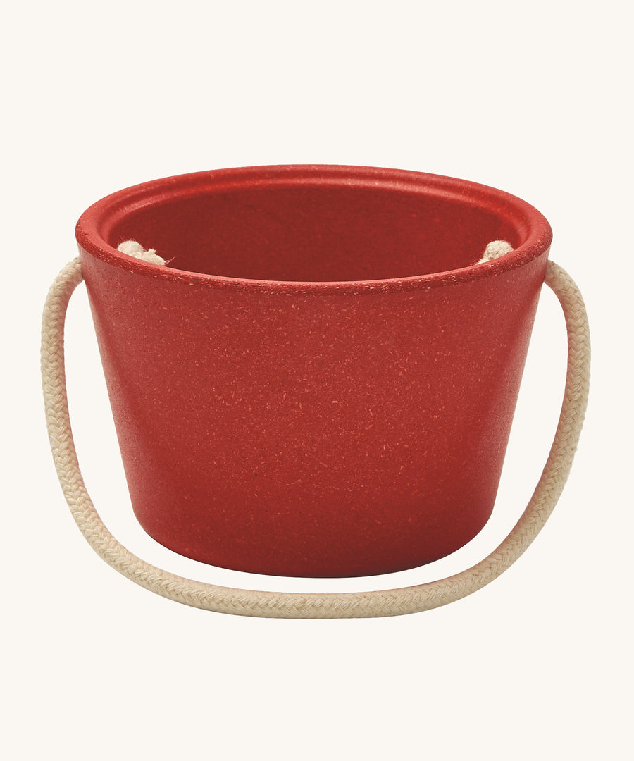 The side of the PlanToys red bucket, from the Sand Play set showing the beige rope handle, on a cream background