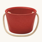 The side of the PlanToys red bucket, from the Sand Play set showing the beige rope handle, on a cream background