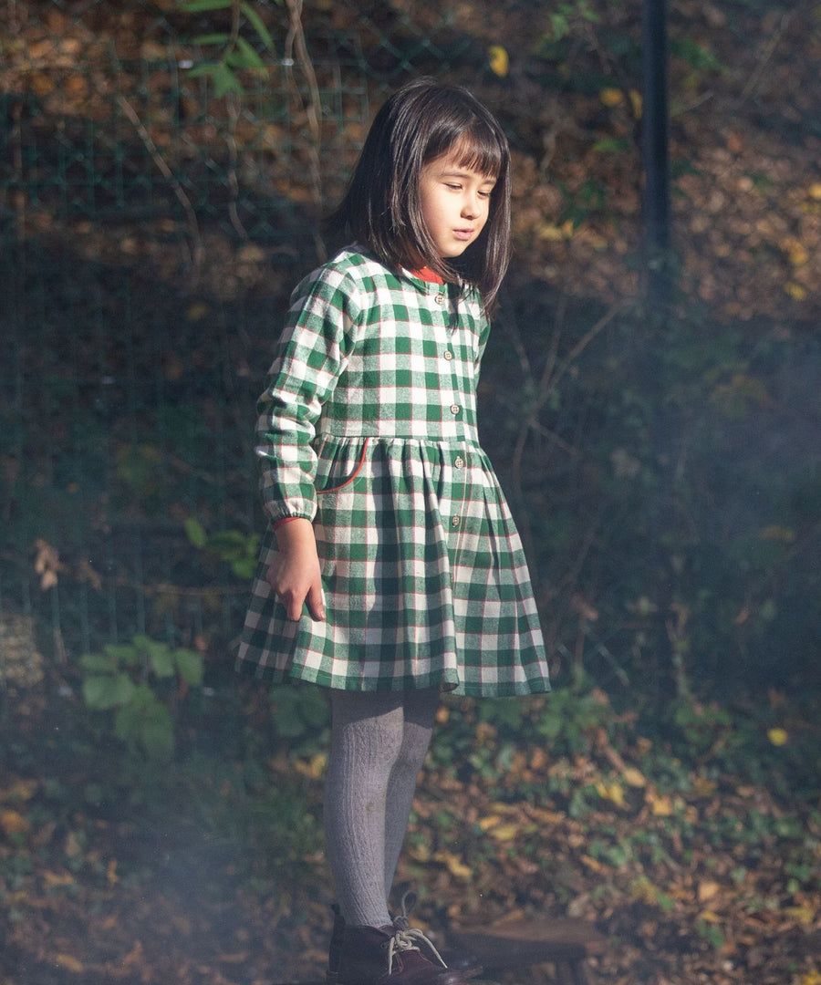 A closer look at the Little Green Radicals Check Pocket Dress - Fern Green, being worn by a child who is stood in the woods and is ready to play with the crunchy autumn leaves