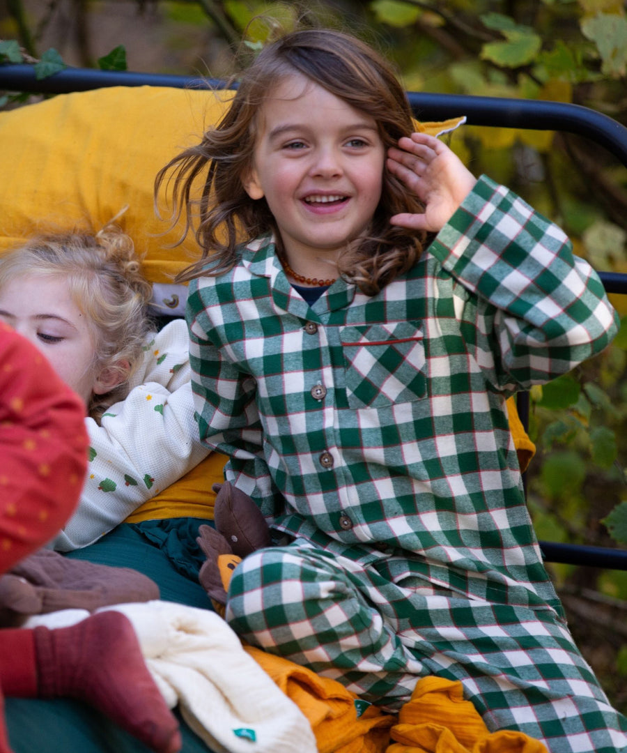 A child sitting on a bed outside with other children, having fun and wearing the Little Green Radicals Classic Check Pyjamas in Fern Green. The image shows a closer look at the check detailing on the pyamas