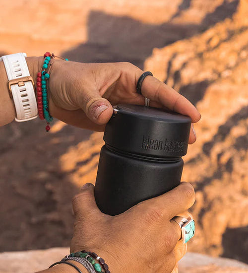 A person twisting the Klean Kanteen Wide Loop Cap - With Bale, in black, with a mountain background