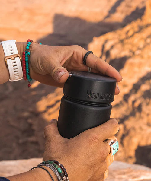 A person twisting the Klean Kanteen Wide Loop Cap - With Bale, in black, with a mountain background