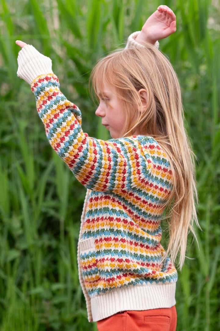 A girl wearing a rainbow stripe knitted cardigan from Little Green Radicals in a forest to represent Little Green Radicals knitwear at Babipur.