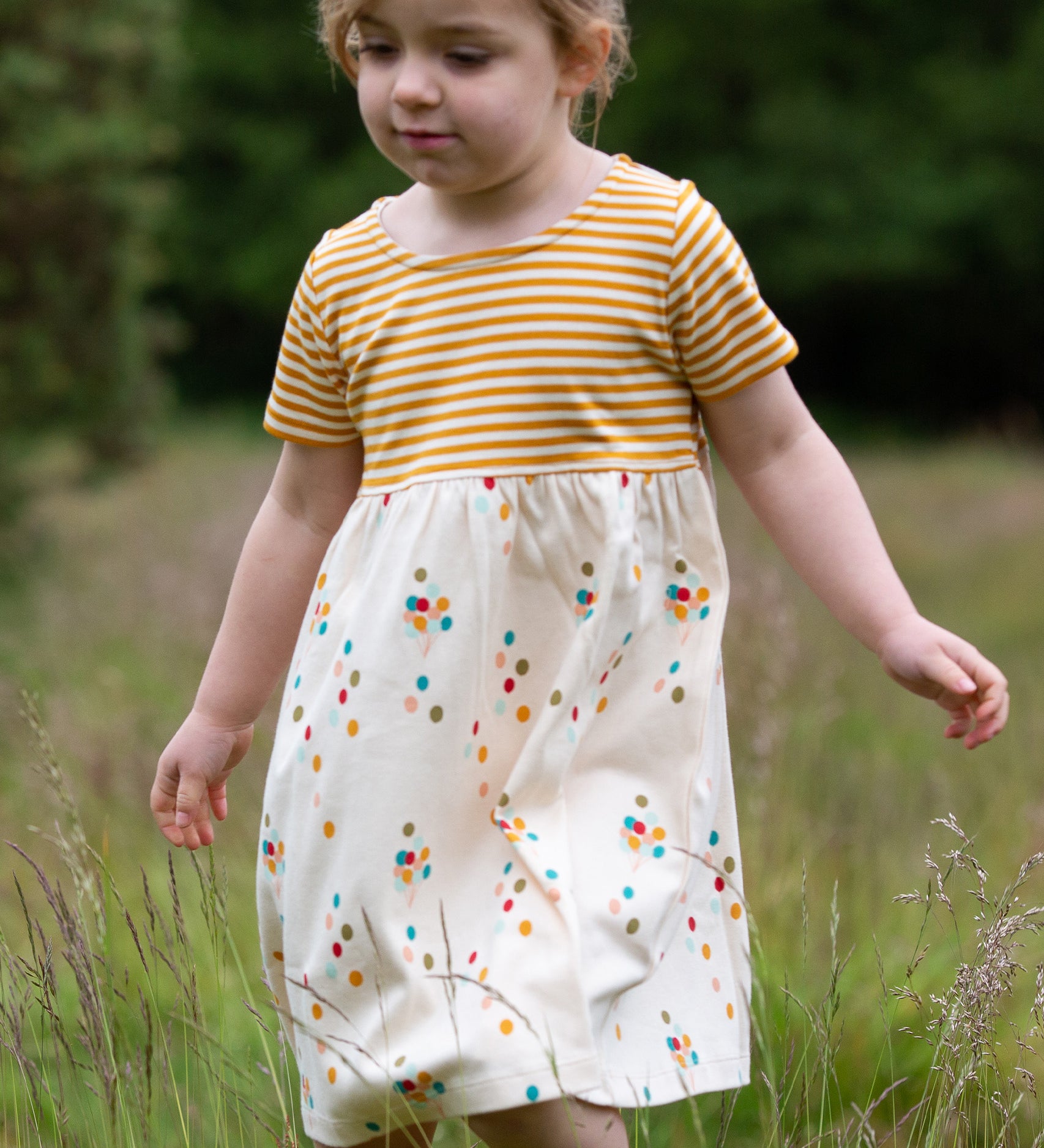 A child playing in a grassy field wearing the LGR Rainbow Balloons Easy Peasy Summer Dress