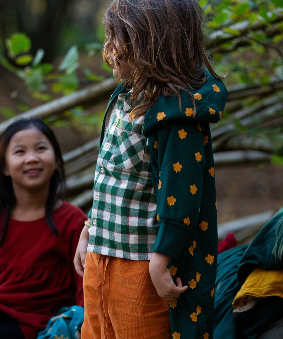 Children playing outside in a forest, and have made a den to play in. The child at the front is wearing the Little Green Radicals Check Long Sleeve Shirt - Fern Green, and a green and yellow cape on top