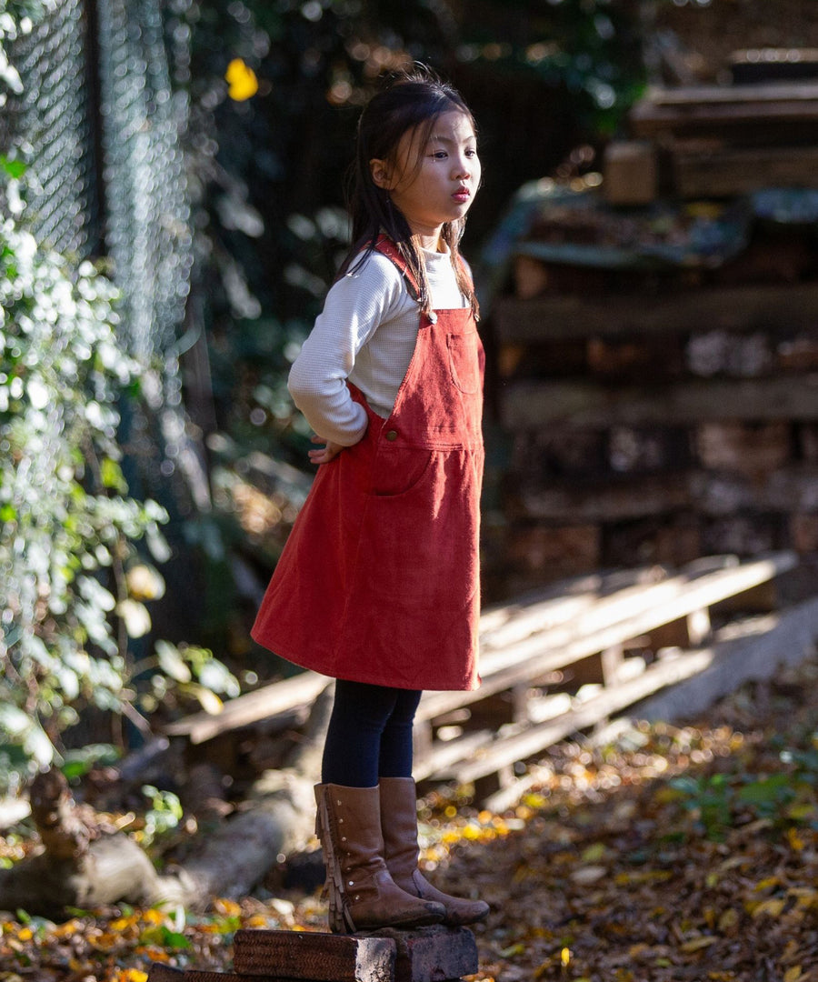 A child stood outside with their hands behind their back, looking afar, wearing the LGR Classic Pinafore Dress - Hazelnut