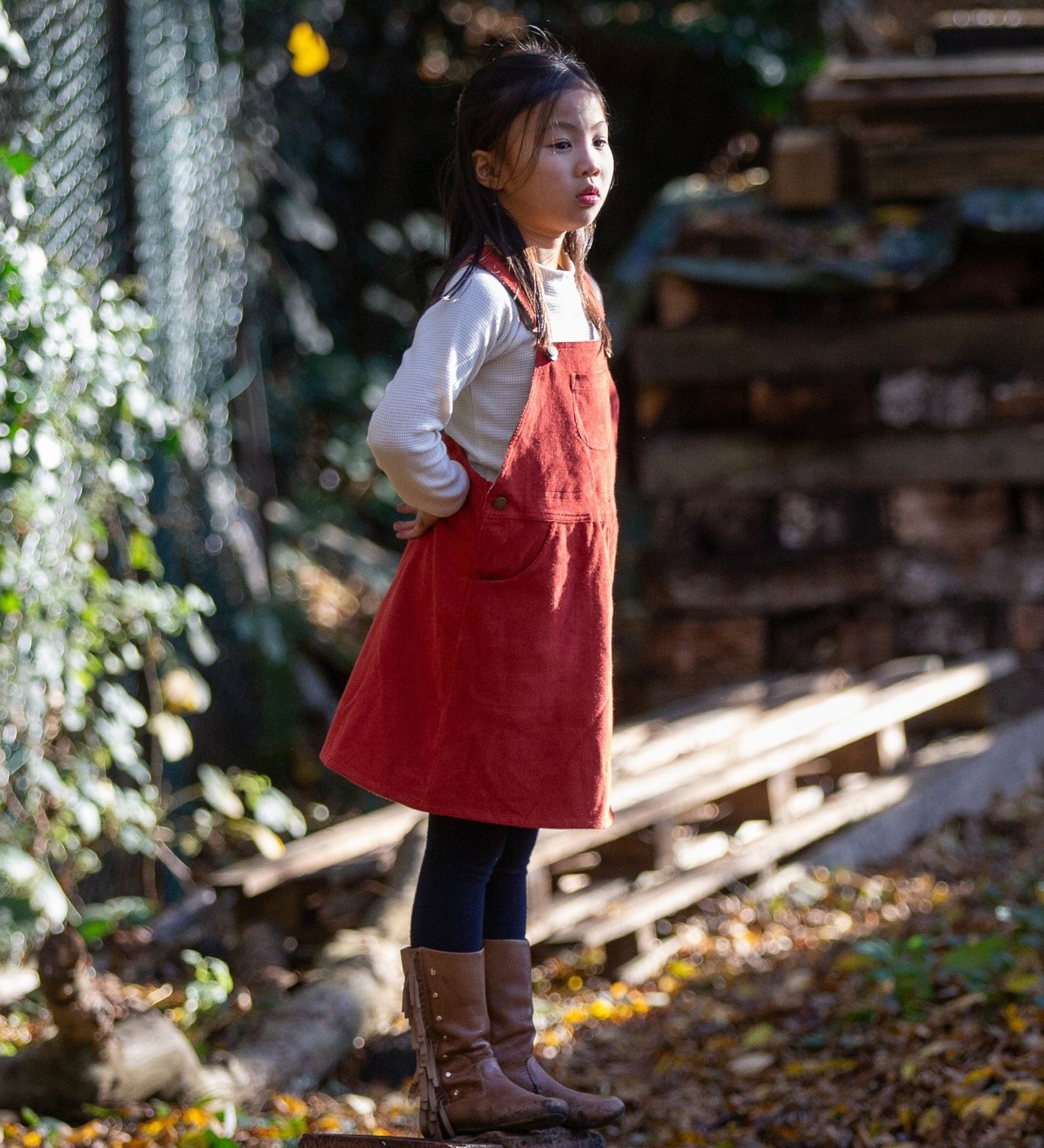 A child stood outside with their hands behind their back, looking afar, wearing the LGR Classic Pinafore Dress - Hazelnut