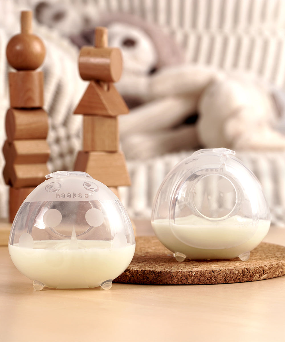 The 40ml Haakaa Ladybug Silicone Breast Milk Collector, filled with milk and stood on a light brown table. The left milk collector shows the ladybird print, and the right milk collector shows the nipple hole on the back