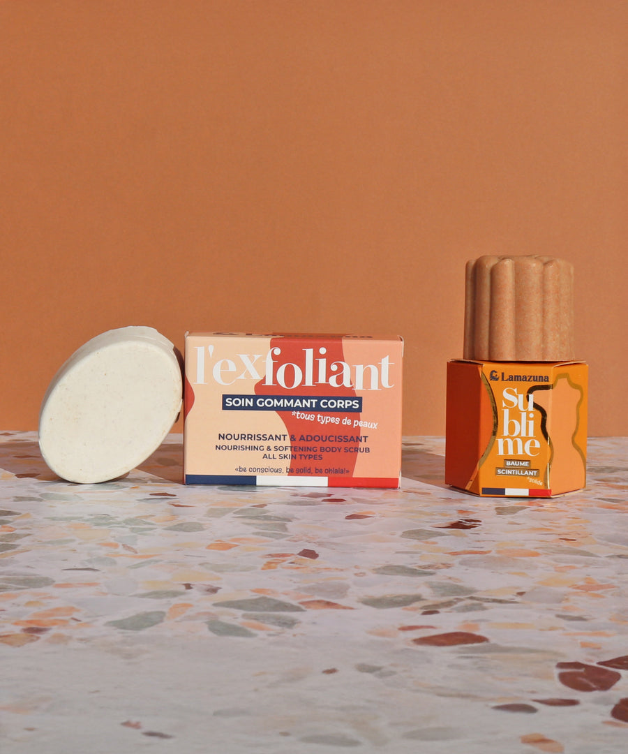 The Lamazuna Solid Exfoliant Bar, and the Lamazuna Sublime Shimmer bar stood next to their packaging boxes, on an orange background and a colourful marble bathroom work surface