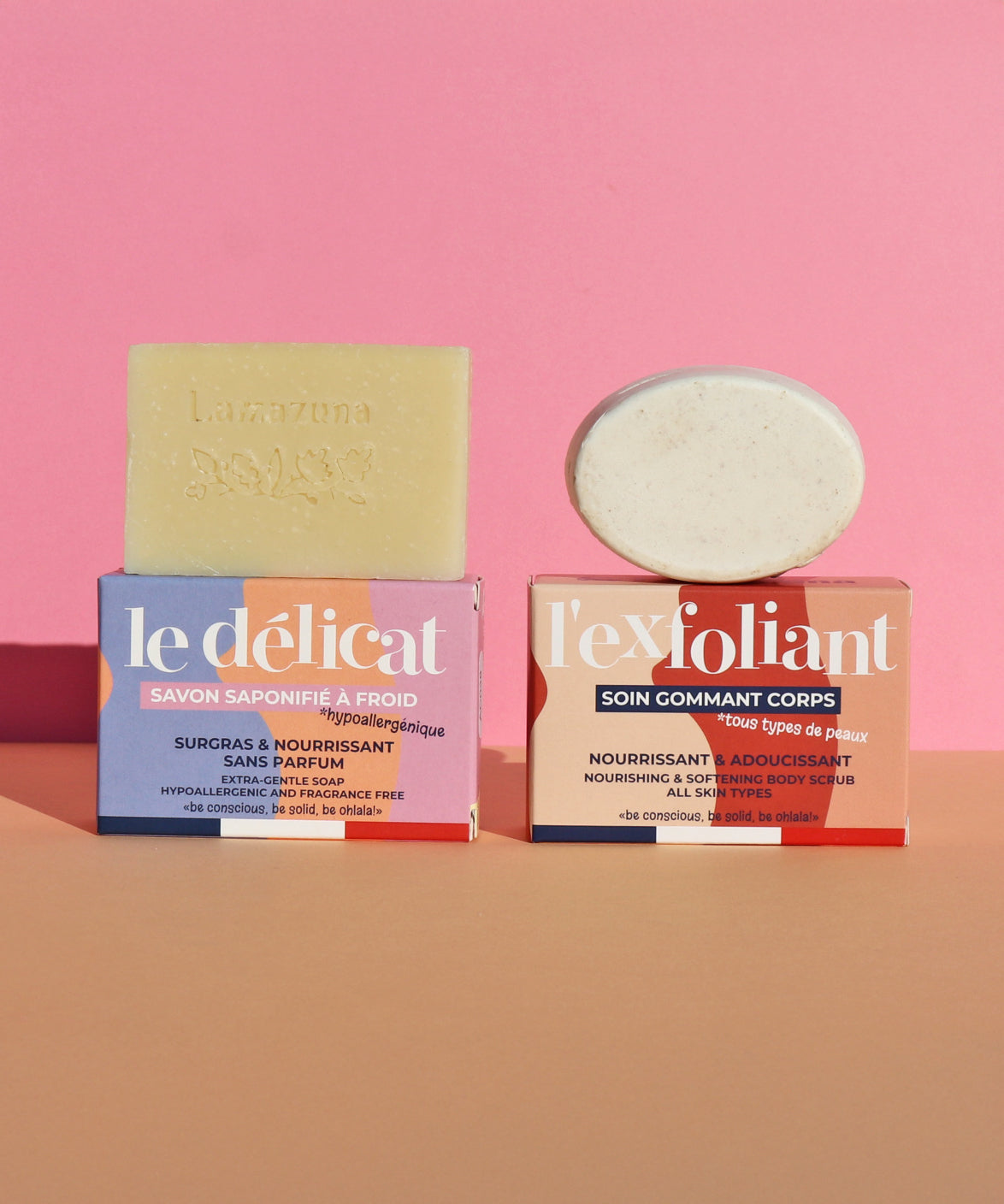 The Lamazuna Solid Exfoliant Bar and the Delicate Gentle Soap Bar, stood on top of their packaging boxes, on a pink and peach background