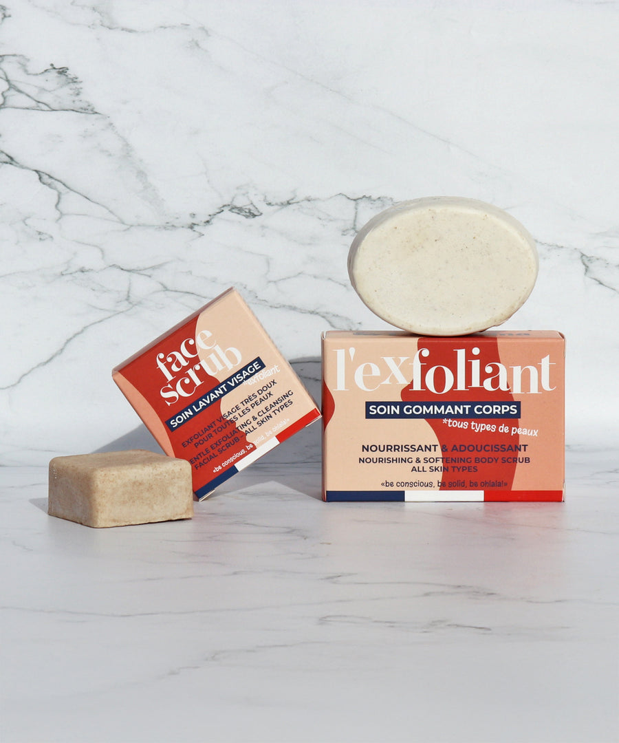 The Lamazuna Solid Face Scrub Bar and the Lamazuna Exfoliating Body Scrub Bar next to each other with their packaging boxes, on a white and grey marbled background