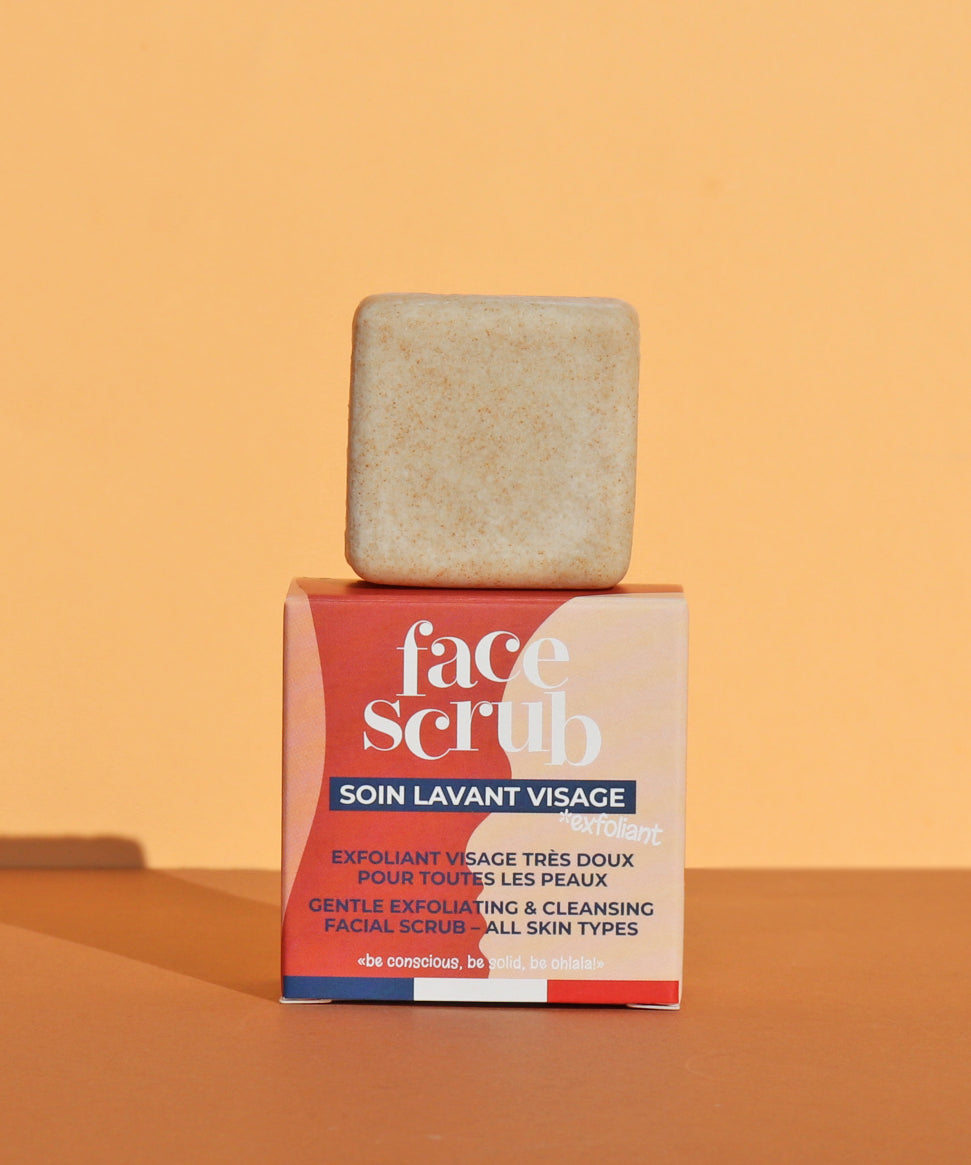 The Lamazuna Solid Face Scrub Bar stood on top of its packaging box, on a yellow and orange background