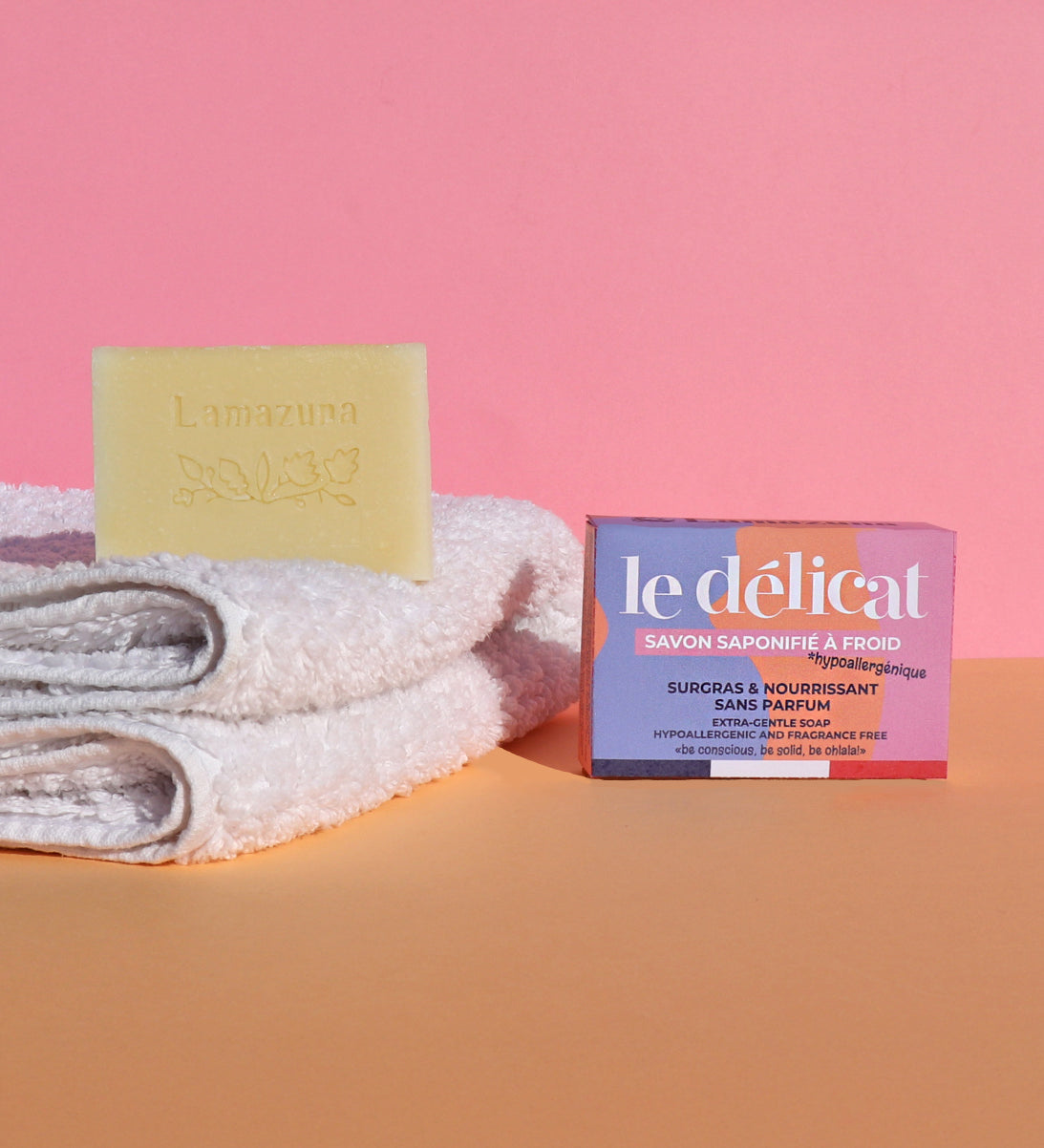 The Lamazuna Sensitive Skin Soap Bar is stood on top of some folded white fluffy towels, with the packaging box text to the towels, on a pink and orange background