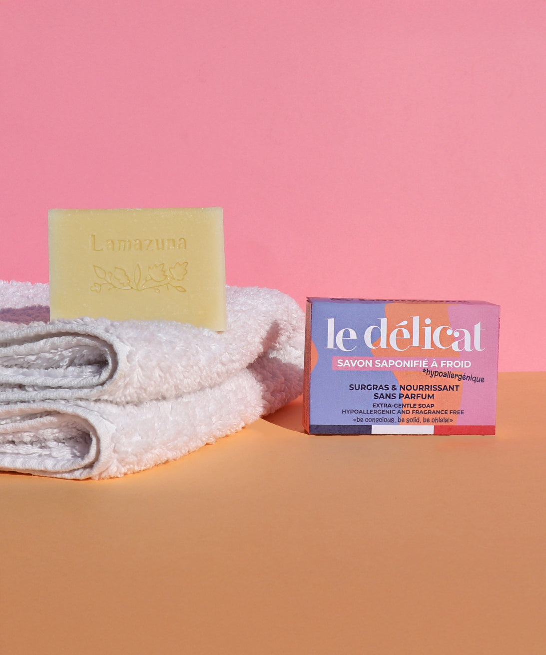 The Lamazuna Sensitive Skin Soap Bar is stood on top of some folded white fluffy towels, with the packaging box text to the towels, on a pink and orange background