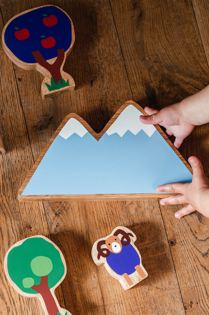 Childs hands reaching out for Lanka Kade x Babipur exclusive wooden Moel figure, a grey/blue mountain with snowy caps, over a dark wooden floor. There are 3 figures scattered on the floor, a purple tree with red apples, a tall green tree and a purple bodied mountain goat with brown horns.