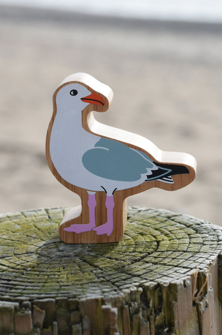 Lanka Kade wooden seagull figure perched on a wooden stump on a sandy beach. The seagull has an orange beak, white body, grey wings and two pink legs and feet.