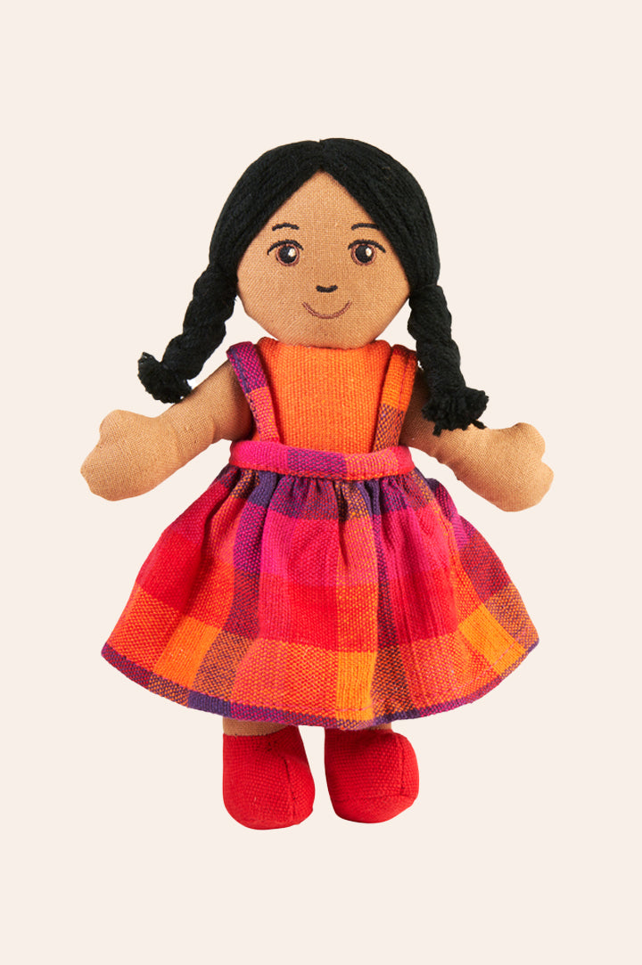 Lanka Kade fair trade rag doll girl with brown skin and plaited black hair. She has brown eyes, black eyebrows and a happy smile. She's wearing a pink and orange woven cotton pinafore dress, red shoes and an orange top.